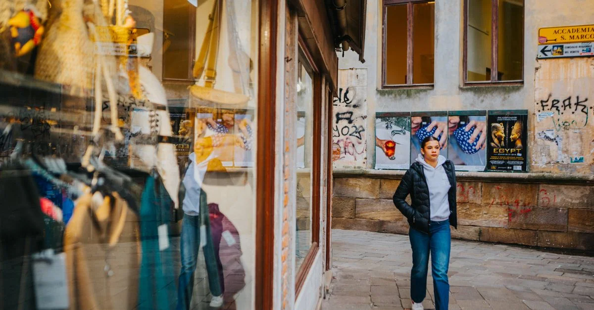 Une jeune femme en tenue décontractée passe devant une vitrine dans une ruelle européenne avec des graffitis et des affiches.