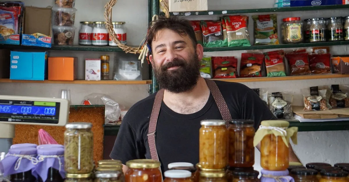Un commerçant affable se tient derrière des étagères remplies de conserves artisanales.