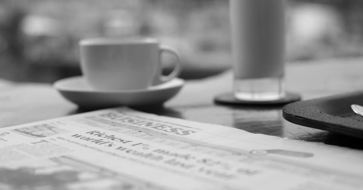 Élégante photo en noir et blanc d'une tasse de café, d'un jus de fruits et d'un journal sur une table dans un cadre serein.