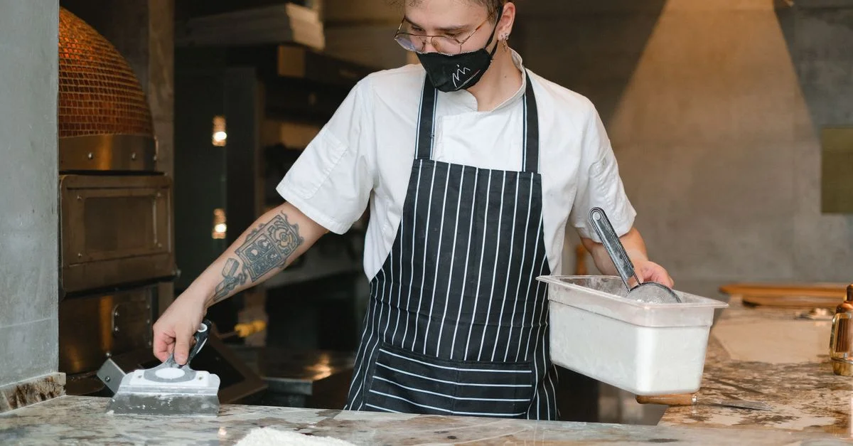 Chef portant un masque et un tablier préparant de la pâte dans un environnement de cuisine professionnelle.